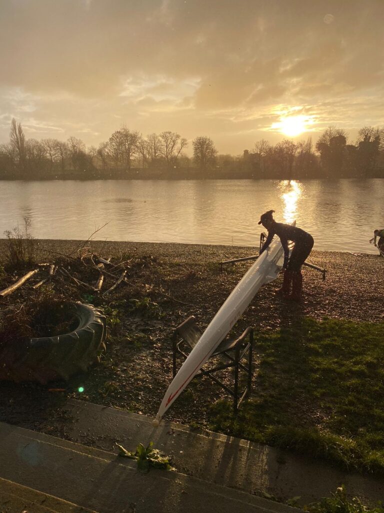 A female rower picking up a single scull rowing boat on a bank by the river Thames at sunset